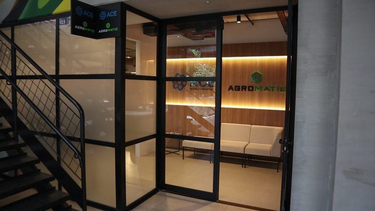 Entrada envidraçada do escritório da Agromatic e ACE, com recepção em madeira ao fundo