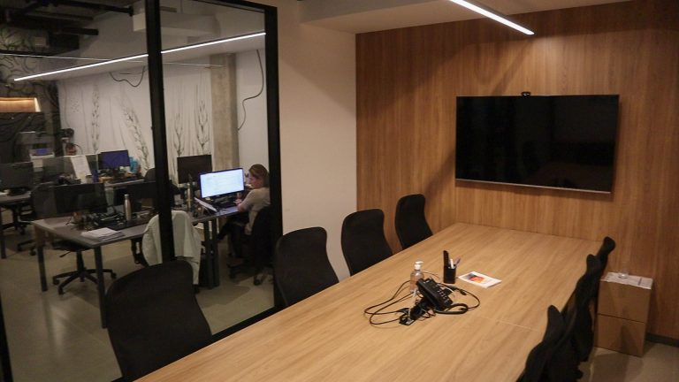 Sala de reunião da Agromatic com mesa de madeira e TV, escritório visível pela divisória de vidro